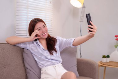 20 'li yaşlarda neşeli bir Asyalı kadın rahat evinde selfie çekiyor. Gülümsüyor ve rahat görünüyor. Kanepede otururken, içeride hoş bir anı yakalıyor..