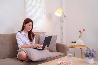 Çekici genç kadın (20-30 'lu yaşlar) kanepede dinlenirken dizüstü bilgisayarla çalışıyor. Merkez ofis doğal ışıkla donatılmış. Rahat ve işine odaklanmış..