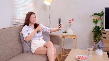 Neşeli bir Asyalı kadın (20-30 yaş) bir kanepede oturur ve akıllı telefonuyla selfie çeker. Ev içi arka plan. Sosyal medya, iletişim ve modern yaşam için yaşam tarzı kavramı.