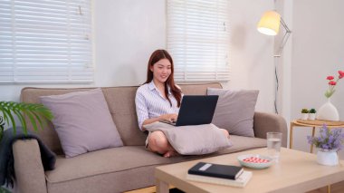 20 'li yaşlarda genç bir kadın evdeki bir kanepede oturuyor ve dizüstü bilgisayarı üzerinde çalışıyor. Odaklanmış ve rahatlamış görünüyor. Uzak çalışma, yaşam tarzı ve teknoloji hakkındaki blog ve makaleler için mükemmel.