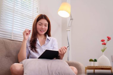 Yirmili yaşların ortasında Asyalı bir kadın evdeki koltukta rahat bir şekilde oturur ve kalemle deftere yazarken gülümser. Doğal ışıkla sakin ve rahat bir ortam.