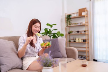 20 'li yaşlarda genç ve güzel bir kadın kanepesinde taze salata yiyor ve sağlıklı bir yaşam tarzını destekliyor. Diyet, sağlık ve ev yemekleri hakkındaki makaleler için mükemmel..