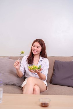 20 'li yaşlarda genç bir kadın evde taze salata yiyor. Kanepede rahatça oturup sağlıklı yemeğinin tadını çıkarıyor. Bu görüntü rahatlama ve sağlıklı beslenme anını yakalar..