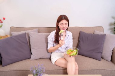 20 'li yaşlarda genç bir kadın taze salata yerken koltukta dinleniyor. Günlük giysiler giyiyor ve sıcacık bir evde sağlıklı yemeğinin tadını çıkarıyor gibi görünüyor..