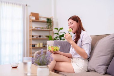 20 'li yaşlarda genç bir kadın koltuğunda taze ve sağlıklı bir salata yiyor. Bu görüntü rahat, sağlığa duyarlı bir yaşam tarzı yakalıyor. Sağlık, diyet ya da yaşam tarzı içeriği için mükemmel.