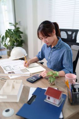 Genç Asyalı kadın mimar yeni ev tasarımı projesi için bina masraflarını hesaplıyor. İnşaatın planlarına ve mühendislik detaylarına bakıyor. Çalışan Asyalı kadın mimar.