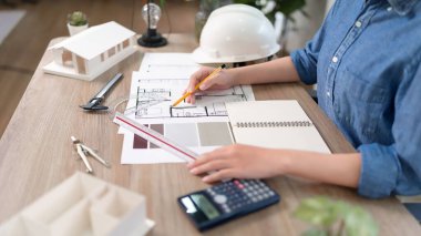 Genç Asyalı bir kadının elleri ahşap bir masanın üzerine bir mimar çizimi yaparken görülüyor. Mimar bir inşaat planı oluşturmak için planlar, araçlar ve renk örnekleri içerir.