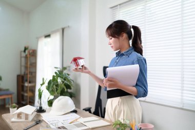 Yetişkin bir Asyalı kadın mimar bir ev modelini inceliyor ve inşaat planlama ve emlak projesi geliştirme konusunda kendine güvenen bir tasarım planı hazırlıyor. Projeyi ve ev modelinin de aynı hizada olmasını sağlayacak planı inceliyor.