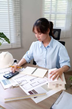 Genç Asyalı kadın mimar hesap makinesi ve ev modeliyle bina tasarım sürecine odaklanan bir tasarım üzerinde çalışıyor. Mimar planları dikkatlice gözden geçirir. Mimari, tasarım ve inşaat görselleri için mükemmel.