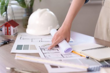 Mimarın Asyalı genç eli ev inşaatı projesinde çalışıyor. Taslak ve güvenlik kaskıyla birlikte. Ev inşaatı projesinde mimari tasarım ve inşaat projesi için planlama yer alıyor..