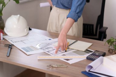 Asyalı mimar bir kadının eli masanın üzerindeki taslağı işaret ediyor. Mimar bir ev inşaatı projesinin tasarımını analiz ediyor. Aletler ve malzemeler, mimar ev planının gelişimini tasarlarken masanın dört bir yanına dağılmış durumda. Kemer.