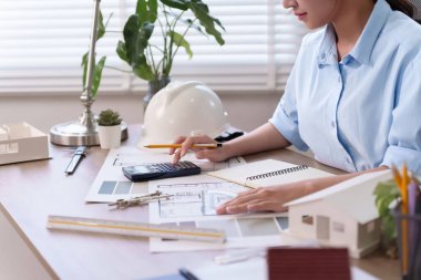 Genç Asyalı kadın mimar titizlikle hesap makinesiyle inşaat planları üzerinde çalışıyor. Proje başarısı için tasarım yapıyor. Bu resim proje başarısı ile mimari tasarımın detaylarını ve hassasiyetini gösteriyor.