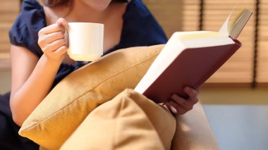 Asyalı bir kadın elinde bir fincan kahve tutarken kitap okuyarak evde boş zamanlarının tadını çıkarıyor. Ellerinde kitap ve rahat bir yastıkla rahatlıyor..