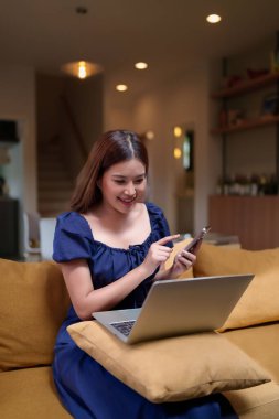 Evdeki kanepede oturan genç Asyalı kadın. Telefonunu kullanıyor ve dizüstü bilgisayarı var. Gülümsüyor ve mutlu görünüyor. Modern yaşam tarzının tadını çıkar.