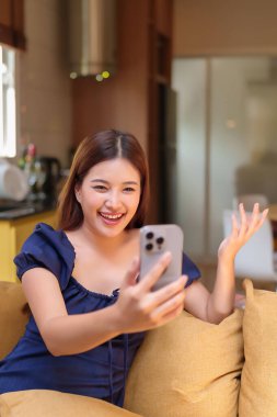 Neşeli yetişkin Asyalı kadın akıllı telefonunu evde video görüşmesi için kullanıyor. Kanepede dinleniyor, gülümsüyor ve başkalarıyla konuşurken el sallıyor..
