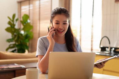 Genç bir Asyalı kadın ev ofisinden uzaktan çalışıyor. Sırıtıyor ve elinde bir dosya tutarken akıllı telefonuyla konuşmaktan zevk alıyor..