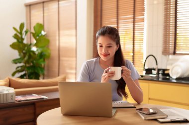 Rahat yetişkin Asyalı kadın, parlak ev ofisinde dizüstü bilgisayarıyla çalışırken kahvesini yudumluyor. Bu modern çalışma alanı düzeniyle üretken ve rahat olun.