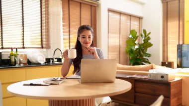 Bu resim, evden dizüstü bilgisayarla çalışırken kahvesini yudumlayan genç bir Asyalı kadını gösteriyor. Çalışma alanı mutfak alanını birleştiriyor. Uzaktan kumandalı, esnek bir yaşam tarzı sağlıyor..