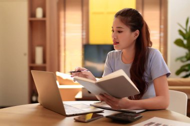 Genç Asyalı kadın evde çalışırken bilgisayar ve kitap okuyarak ve evde oturarak teknoloji ve rahatlıkla eğitim alarak verimliliği artırıyor.