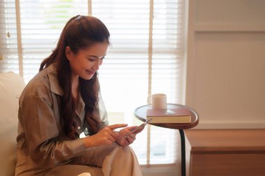 Sıradan kıyafetler giyen Asyalı bir kadın akıllı telefon kullanarak evde dinleniyor. Telefonuna gülümseyerek bakıyor, iletişim için modern bir bağlantı sergiliyor..