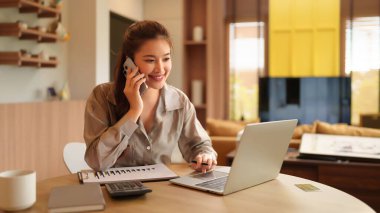 Genç Asyalı kadın evinde bilgisayarını kullanarak ve akıllı telefonuyla konuşarak mutlu bir şekilde çalışıyor. Ekrana bakıyor ve iş yaparken gülümsüyor ve uzaktan iletişimi yönetiyor.