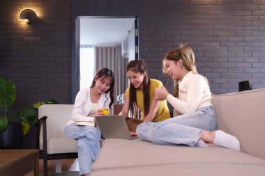 Günlük giysiler içinde üç mutlu Asyalı kadın evdeki kanepede dizüstü bilgisayar kullanarak online alışveriş için işbirliği yapıyorlar. Birlikte kredi kartı kullanarak ticaretten zevk alan genç arkadaşlar..