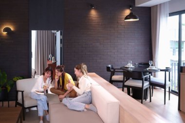 Genç görünen üç Asyalı kadın modern bir dairede dizüstü bilgisayarda birlikte çalışıyorlar. Kanepede rahatça oturuyorlar, fikirleri paylaşıyorlar ve bir projede işbirliği yapıyorlar. Yaşam alanı parlak ve şık bir iç tasarımı var. Mükemmel resim...