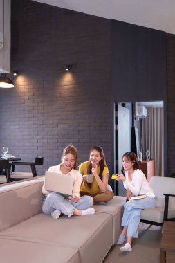 Üç Asyalı bayan arkadaş evde boş zamanın tadını çıkarıyorlar. Biri dizüstü bilgisayar kullanıyor, diğeri kredi kartı tutuyor, diğeri de bardakla. Birlikte rahatlıyorlar, internetten alışveriş yapabilirler..