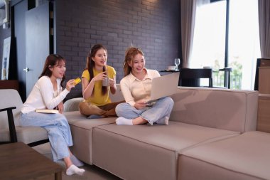 Üç Asyalı genç kadından oluşan bir grup evdeki rahat bir kanepede dinleniyor. Bir tanesi bilgisayarla online alışveriş yapıyor ve bu deneyimi arkadaşlarıyla paylaşıyor. Birlikte mutlu bir şekilde kahve içiyorlar..