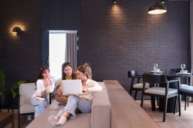 Üç genç Asyalı kadın şık bir oturma odası ortamında online alışveriş yapmayı seviyorlar. Diz üstü bilgisayar ve kredi kartıyla ekomünikasyon işi yapıyor. İçerideki konforlu oturma odasına odaklan..