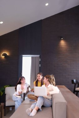 Üç genç Asyalı kadın modern bir kapalı mekanda. Biri dizüstü bilgisayar kullanıyor ve kredi kartı tutuyor. Diğeri pembe kahve fincanı tutuyor. Birlikte dinleniyorlar, sosyalleşiyorlar, çalışıyorlar, internette geziniyorlar ya da alışveriş yapıyorlar. Mutlu anlar, arkadaşlar..