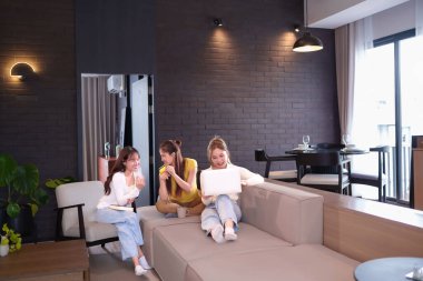 Üç genç Asyalı kadın modern bir oturma odasında dinleniyor, biri dizüstü bilgisayar kullanıyor, diğeri kredi kartı tutuyor, ve biri de kitabı olan, birlikte zaman geçirip internetten alışveriş yapan veya sosyalleşen biri..