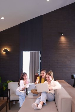 Üç genç Asyalı kadın, modern bir oturma odasında dizüstü bilgisayarda birlikte çalışıyor ve kahve içerek fikirlerini paylaşıyorlar. Bu takım çalışması dijital teknolojiyi kullanarak başarıyı gösteriyor..