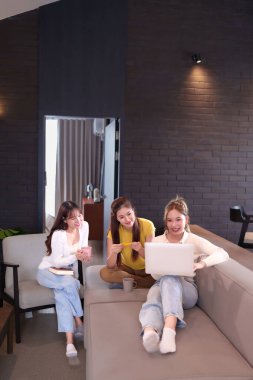 Üç Asyalı genç kadın rahat bir oturma odasında oturuyorlar ve birlikte dizüstü bilgisayardan alışveriş yapıyorlar. Onlar evlerinde modern bir yaşam tarzını paylaşan iyi arkadaşlardır..
