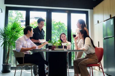 Yetişkin Asyalı arkadaşlar ve meslektaşlar yemek ve içeceklerle gündelik bir toplantı yapıyorlar. Tatil zamanı modern ofis ortamında birlikte gülüyorlar..