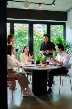 Asyalı genç profesyonellerden oluşan çeşitli bir grup modern bir ofisteki masanın etrafında iş birliği yapıyor, fikir alışverişinde bulunuyor ve proje başarısını geliştirmek için birlikte ara veriyorlar.