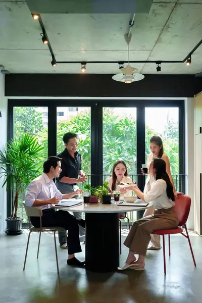 Bir grup Asyalı profesyonel yetişkin modern, parlak bir ofis alanında yemek yiyor ve yuvarlak bir masanın etrafında sohbet ediyorlar. Yiyecek ve içecekleri paylaşıyorlar.