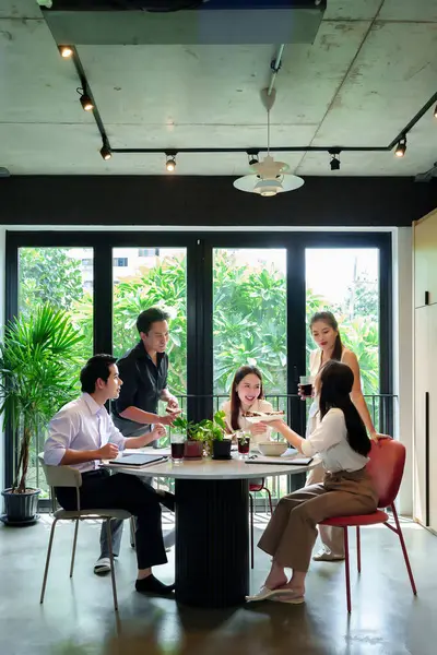 Modern bir ofisteki bir grup Asyalı yetişkin, bir masa etrafında içki ve abur cubur paylaşıp takım çalışmalarını tartışarak iş birliği yapıyor.