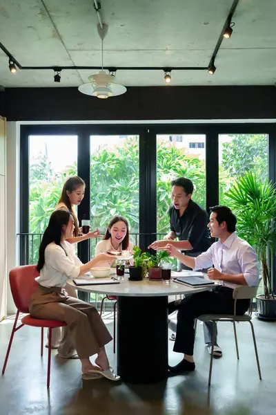 Bir grup genç Asyalı iş adamı öğle yemeği için parlak bir ofiste bir masanın etrafında toplanıyorlar. Yemeklerini paylaşıyorlar, iletişim kuruyorlar ve toplantılarında işbirliği yapıyorlar..