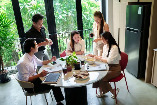 Bir grup Asyalı yetişkin, yuvarlak bir masada iş toplantısı yapıyorlar. Modern bir ofiste yemek yiyip içki içiyorlar. Fikirler üretmek ve stratejiler üzerinde işbirliği yapmak.