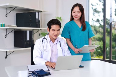 Asyalı yetişkin doktor ve hemşire hasta kayıtlarını incelemek için bir dizüstü bilgisayar kullanarak modern bir klinikte iş birliği yapıyorlar. Sağlık hizmetlerine tıbbi uzmanlık ve ekip çalışması sağlıyorlar..