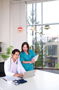 Meslektaşı hemşire ile dizüstü bilgisayar kullanan genç Asyalı yetişkin doktor, parlak modern klinikte tıbbi kayıtları tutuyor. Sağlık hizmetleri ekip çalışması konsepti.