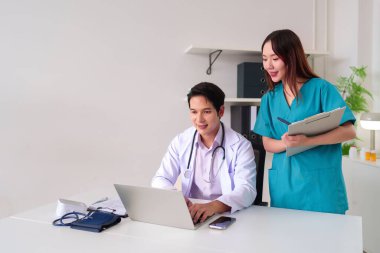 Yetişkin bir Asyalı doktor dizüstü bilgisayar kullanırken yetişkin bir Asyalı hemşire dosya tutuyor. Hasta kayıtlarını ve sağlık danışmanlığını görüşmek için bir klinikte birlikte çalışıyorlar..