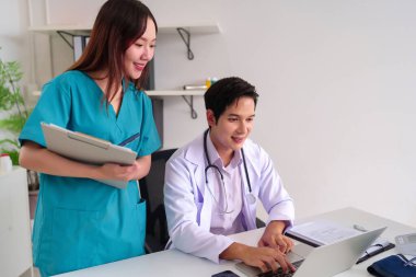 Yetişkin Asyalı doktor hasta verileri için bir dizüstü bilgisayar kullanıyor. Hemşire klinikte bir sağlık dosyası tutuyor. Modern bir ofis ortamında hasta bakımında işbirliği yapıyorlar..