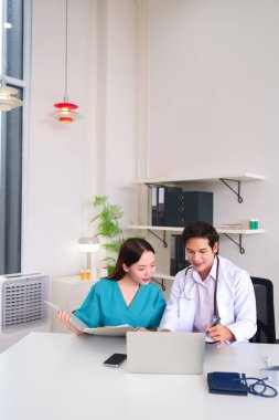 İki Asyalı sağlık uzmanı yetişkin bir doktor ve hemşire hastane masasında işbirliği yaparak en iyi hasta bakımı uygulamaları için gerekli belgeleri ve dizüstü bilgisayarı inceliyorlar.