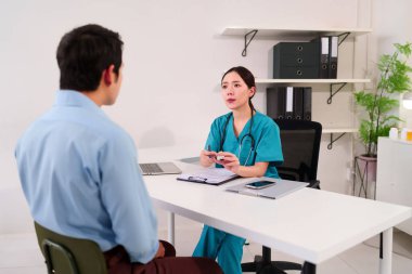 Ofisindeki Asyalı kadın doktor, yetişkin bir erkek hastayla sağlık sorunları ve tedavi seçenekleri hakkında görüşüyor. Sağlık ve refah hizmetlerine odaklanıyorlar..