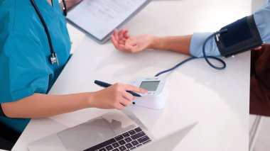 Önlüklü bir Asyalı hemşire yetişkin bir hastanın sağlığını incelemek için dijital tansiyon monitörü kullanıyor. Hemşire not alıyor. Klinikteki masada dizüstü bilgisayarı olan bir kalem kullanıyor.