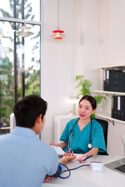 Asyalı bir kadın doktor, tıbbi konsültasyon sırasında yetişkin bir erkeğin tansiyonunu ölçüyor. Daha iyi bir tedavi için modern bir klinikte sağlığı tartışıyorlar.