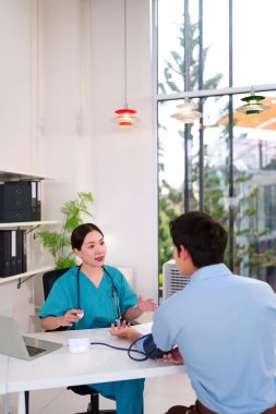 Yeşil önlüklü Asyalı kadın doktor, sağlık ve tıbbi tedavi için klinikteki sfenometreyle Asyalı erkek hastanın kan basıncını ölçüyor..
