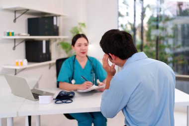 Yeşil üniformalı Asyalı yetişkin kadın doktor klinikte baş ağrısı çeken erkek hastayla görüşüyor. Adam hastayken doktor notlar alıyor.